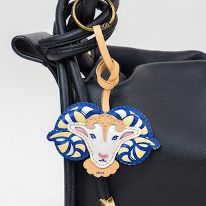 May include: Aries zodiac sign keychain. The keychain features a ram design with blue, yellow, and white accents. The keychain is attached to a leather strap and a gold-colored ring. The word "ARIES" is printed on the bottom of the keychain.