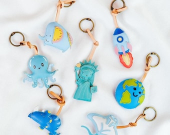 May include: Collection of keychains featuring various designs, including an elephant, octopus, rocket, Statue of Liberty, globe, dinosaur, and stingray. Each keychain has a leather strap and a metal ring, perfect for keys or bags.
