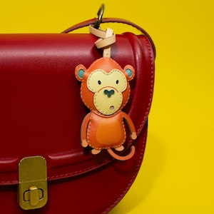 May include: A leather monkey keychain in shades of orange, tan, and light blue, attached to a red handbag. The keychain features a stitched design with a surprised expression. The background is a bright yellow.