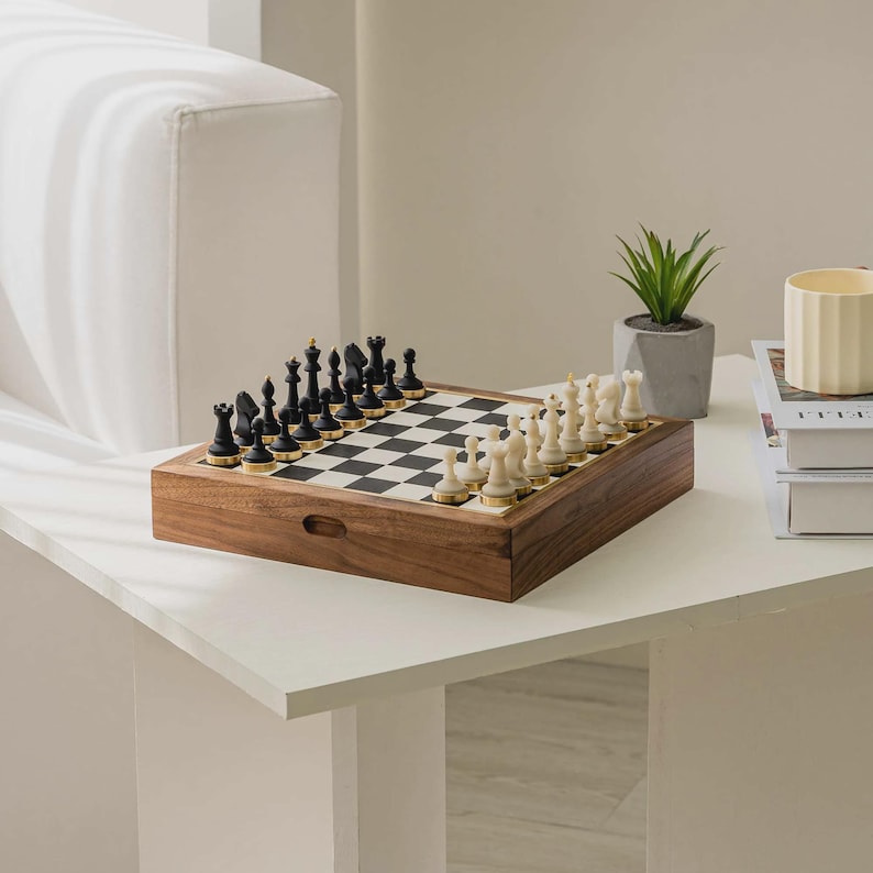 May include: A wooden chess set with black and white pieces arranged on a checkered board. The chess pieces are black and cream-colored. The wooden box has a drawer. A small potted plant and a candle are in the background.