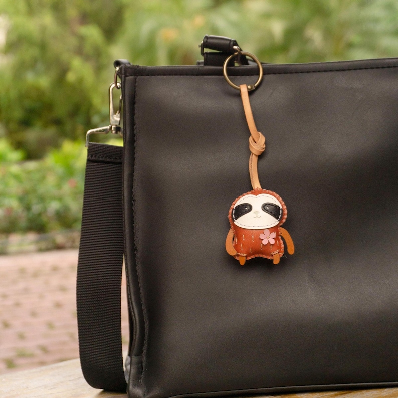 May include: A brown leather sloth keychain with a pink flower accent hangs from a black purse. The sloth has black eyes and a white face. The keychain is attached to a gold-colored ring.
