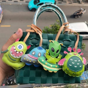 Könnte beinhalten: Eine grüne gewebte Tasche mit vier bunten Schlüsselanhängern zum Thema Außerirdische. Die Schlüsselanhänger zeigen einen grünen Außerirdischen mit einem Raumschiff, ein grünes Monster mit rosa Akzenten, ein blaues Raumschiff und einen grünen Außerirdischen mit einem Raumschiff.