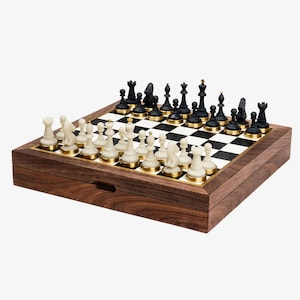 May include: A wooden chess set with black and white pieces arranged on a checkered board. The chess pieces are black and off-white, with gold-colored accents. The board is set inside a wooden box.