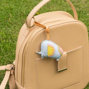 May include: A light blue elephant-shaped keychain with yellow and pink accents hangs from a tan backpack. The keychain is attached to a leather strap. The backpack is tan and has a front pocket.