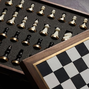 May include: A wooden chess set with a black and white checkered board. The chess pieces are stored in a foam-lined compartment. The pieces are black and white with gold-colored accents.