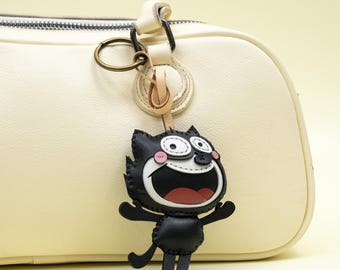 May include: A black cat keychain with a wide open mouth and surprised expression. The keychain is attached to a cream-colored bag with a gold-colored ring and leather strap. The cat has white eyes, pink cheeks, and a red mouth.
