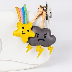 Könnte beinhalten: Eine weiße Ledertasche mit einem silbernen Metallring und einem daran befestigten Anhänger. Der Anhänger zeigt einen gelben Stern mit einem Smiley, einen Regenbogen und eine graue Wolke mit einem bösen Gesicht und Blitzen.