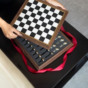 May include: A wooden chess set with a black and white checkered board and a gold-trimmed frame. The chess pieces are black and white with gold accents, nestled in a foam-lined box. A red ribbon is wrapped around the box.
