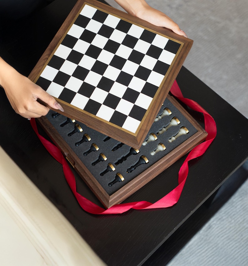 May include: A wooden chess set with a black and white checkered board and a gold-trimmed frame. The chess pieces are black and white with gold accents, nestled in a foam-lined box. A red ribbon is wrapped around the box.