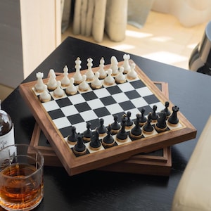 May include: A wooden chess set with black and white checkered board and pieces. The chess pieces are black and white with gold accents. The set is displayed on a black table with a glass of amber liquid and a bottle.