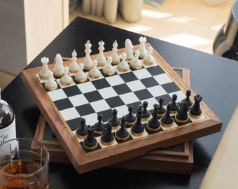 May include: A wooden chess set with black and white checkered board and pieces. The chess pieces are black and white with gold accents. The set is displayed on a black table with a glass of amber liquid and a bottle.
