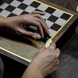 May include: A person's hands working on a wooden chessboard. The board has a gold border and a black and white checkered pattern. A small, light-colored tool is being used to work on the board.