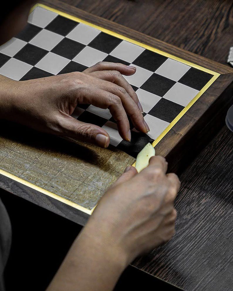 May include: A person's hands working on a wooden chessboard. The board has a gold border and a black and white checkered pattern. A small, light-colored tool is being used to work on the board.
