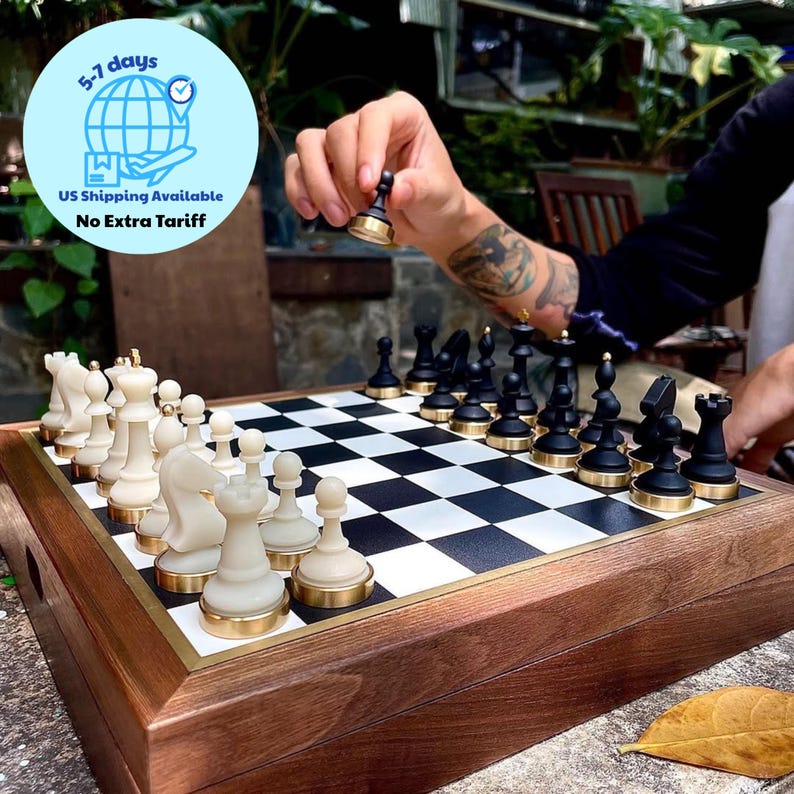 May include: A wooden chess set with black and white pieces. The chess pieces are black and cream-colored with gold-colored bases. The board is black and white with a gold-colored border. The text in the image says "US Shipping Available" and "No Extra Tariff".