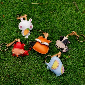 May include: A collection of handmade leather keychains in the shape of animals, including a white bunny holding a carrot, a red bull, an orange fox, a black and white cow, and a blue elephant. Each keychain has a metal ring and leather strap.