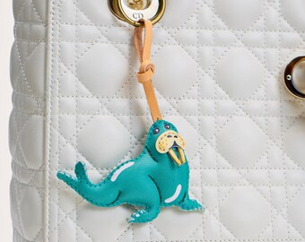 May include: A white quilted handbag with a teal blue walrus charm attached to the top handle. The charm has a tan leather strap and a gold metal clasp.