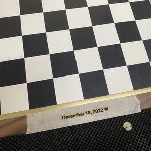 May include: A close-up of a chessboard with alternating black and white squares. The board has a wooden frame with a gold border. The text "December 18, 2022" is visible on a piece of tape adhered to the board.