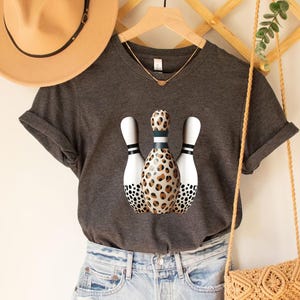 May include: A dark gray t-shirt with a graphic of three bowling pins. The middle bowling pin is covered in a leopard print pattern. The other two bowling pins are white with black polka dots.