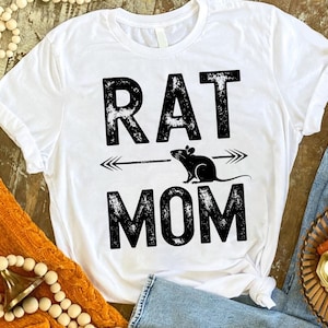 A white t-shirt with the text "RAT MOM" printed on it, along with a graphic of a rat and an arrow. The t-shirt is placed on a wooden surface, surrounded by various items such as beads, a sweater, and a basket.