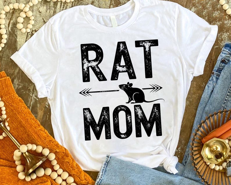 A white t-shirt with the text "RAT MOM" printed on it, along with a graphic of a rat and an arrow. The t-shirt is placed on a wooden surface, surrounded by various items such as beads, a sweater, and a basket.