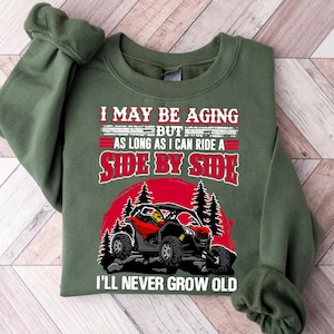 May include: Green sweatshirt with the text "I MAY BE AGING BUT AS LONG AS I CAN RIDE A SIDE BY SIDE I'LL NEVER GROW OLD". The design includes a red and black side-by-side vehicle, a red sun, and pine trees.