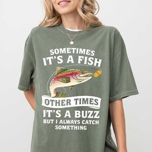May include: Olive green t-shirt with the text "SOMETIMES IT'S A FISH OTHER TIMES IT'S A BUZZ BUT I ALWAYS CATCH SOMETHING". The shirt features a colorful illustration of a fish and a fishing lure.