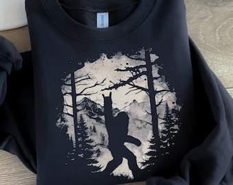 May include: Black sweatshirt with a graphic print of a silhouette of a Bigfoot standing in a forest with mountains in the background.