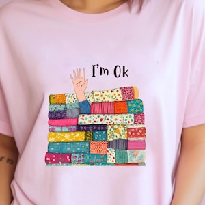 May include: Pink t-shirt with a colorful patchwork design and a hand reaching out from the pile with the text "I'm Ok" above it.