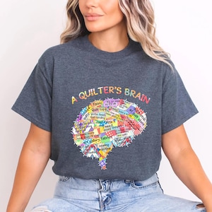 May include: Dark gray t-shirt with the text "A Quilter's Brain" and a colorful brain graphic filled with quilting-related words. The shirt is a comfortable, casual style.