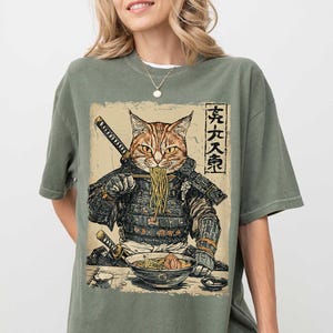 May include: A green t-shirt with a graphic print of a cat dressed as a samurai eating ramen noodles. The Japanese text on the shirt reads "天下一等".
