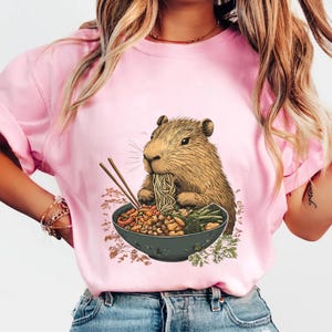 May include: A pink t-shirt with a graphic of a capybara eating noodles from a bowl with chopsticks. The capybara is brown and has a white snout.