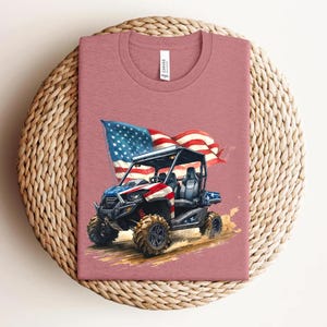 May include: A dusty rose-colored t-shirt featuring a graphic of an off-road vehicle with an American flag waving in the background. The vehicle is painted in red, white, and blue, with black tires and a black roll cage. The shirt has a crew neck.