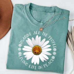 May include: A sage green t-shirt featuring a large white daisy graphic with a yellow center containing a peace sign. The text "IMAGINE ALL THE PEOPLE LIVING LIFE IN PEACE" is printed in a circular white font around the daisy, accented by small flowers.
