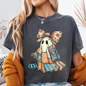 May include: A charcoal gray t-shirt featuring a graphic of a ghost wearing a large floral patterned bow. The ghost is depicted as a patchwork figure in shades of blue, orange, and cream, holding a small blue jack-o'-lantern candy bucket.