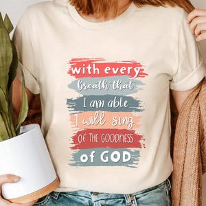 May include: A beige t-shirt with a colorful graphic design that reads "with every breath that I am able I will sing of the goodness of GOD".