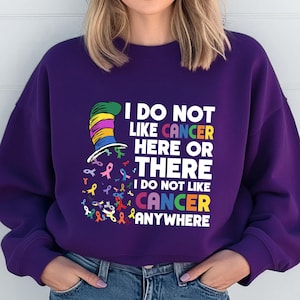 May include: Purple sweatshirt with a colorful graphic of a hat and ribbons. The text on the shirt reads "I do not like cancer here or there I do not like cancer anywhere".