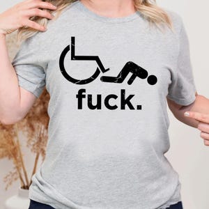 May include: A light gray short-sleeved t-shirt featuring a black graphic of a person in a wheelchair performing a sit-up exercise. Below the graphic, the word "fuck." is printed in a bold, black, sans-serif font. The graphic has a distressed texture.