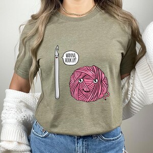 May include: Olive green t-shirt featuring a cartoon crochet hook and a pink yarn ball with a smiling face. The text "WANNA HOOK UP?" is in a speech bubble. The shirt is worn by a person.