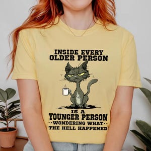 A yellow t-shirt with a cartoon cat and text that reads "Inside every older person is a younger person... wondering what happened".