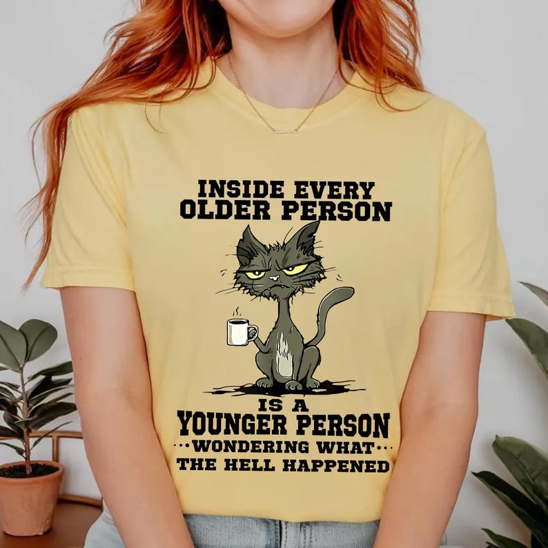 A yellow t-shirt with a cartoon cat and text that reads "Inside every older person is a younger person... wondering what happened".