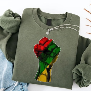 May include: Olive green sweatshirt featuring a raised fist graphic in red, green, and yellow. A silver necklace with a circular pendant is draped across the top. The sweatshirt is paired with blue denim jeans.