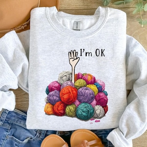May include: A gray sweatshirt with the text "I'm OK" and a cartoon hand sticking out of a pile of colorful yarn.