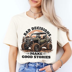 May include: A white t-shirt with a graphic of an off-road vehicle driving through a mountain landscape. The text "Bad Decisions Make Good Stories" is printed on the shirt.