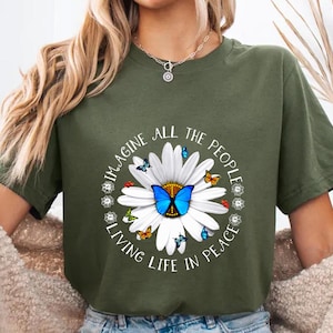 Peut inclure: T-shirt vert olive avec un motif de marguerite et de papillon. La marguerite a un papillon bleu au centre et est entourée de petits papillons. Le texte dit "IMAGINE ALL THE PEOPLE LIVING LIFE IN PEACE."