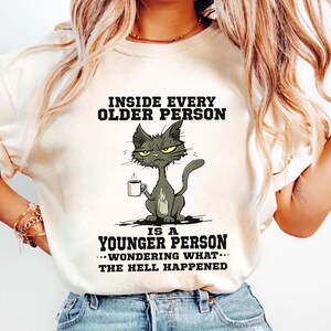 A woman wearing a white t-shirt with a cartoon cat and text that reads "Inside every older person is a younger person... wondering what happened".
