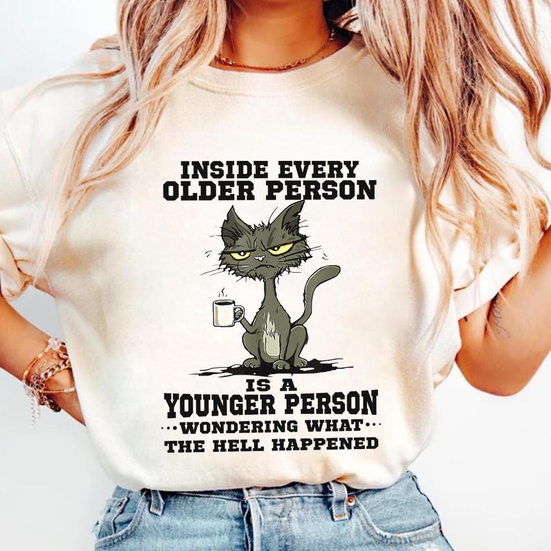 A woman wearing a white t-shirt with a cartoon cat and text that reads "Inside every older person is a younger person... wondering what happened".