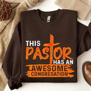 Puede incluir: Sudadera marrón con un gráfico naranja que dice "This Pastor Has An Awesome Congregation" con una cruz detrás de la palabra "Pastor".