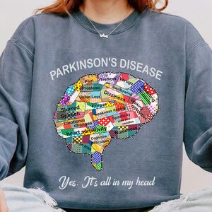 May include: A blue sweatshirt with a brain graphic made of colorful patchwork squares. Each square has a symptom of Parkinson's Disease written on it. The text "PARKINSON'S DISEASE" is at the top of the graphic. The text "Yes. It's all in my head" is at the bottom of the graphic.