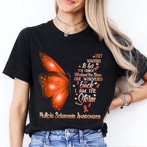 May include: Black t-shirt with an orange butterfly and the text "They whispered to her you cannot withstand the storm she whispered back I am the storm" and an orange ribbon with the text "Multiple Sclerosis Awareness".