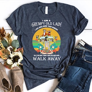 May include: Dark blue t-shirt with the text "I AM A GRUMPY OLD LADY I LOVE DOGS MORE THAN HUMANS". The graphic features a yellow van with dogs and a woman meditating. The text "SO IF YOU DON'T WANT YOUR FEELINGS HURT WALK AWAY" is also included.